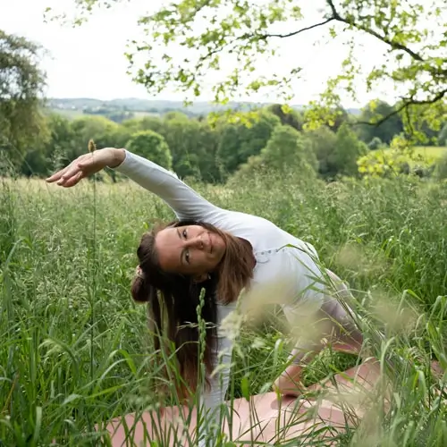yoga-bad-kohlgrub-monika-denca-2 Monika Denca, Yogalehrerin inmitten von hohem Gras, schaut nach vorne und lächelt und macht die Asana - Rockstar Pose Camatkarasana.